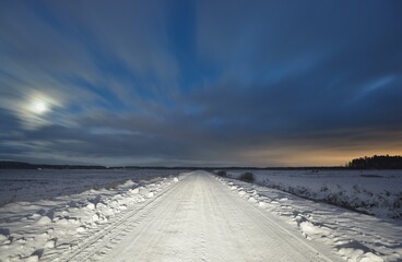 Snow-covered country road through the field and forest at night. Terrific sunset sky, colorful blue...