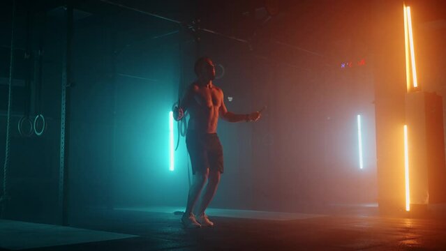 Muscular man with bare torso is jumping using a skipping rope. Male boxer with skipping rope. Fighter doing jump rope exercises in colorful light with smoke in slow motion.