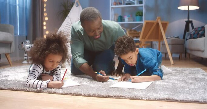 Little african american boy spends weekend with grandparents lying on the floor and drawing with colored pencils.