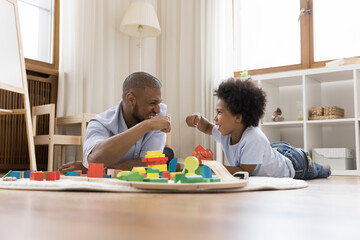 Cheerful African dad and little 6s curly haired son fist bumping making bet start railcars competition, play with wooden rail road lying on warm floor in modern playroom. Family leisure, hobby concept