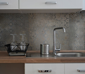 Kitchen sink with tap on wooden counter, texture tiles on the wall, white furniture in modern interior of kitchen.  