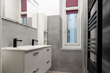 Interior of bathroom with double sink, white furniture, grey tiles and mirror on the wall. Window in modern bathroom.