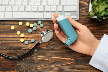 Female doctor holding asthma inhaler, keyboard, pills and stethoscope on wooden background