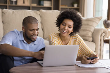 Smiling African couple calculate their earnings and expenses use calculator, pay utility bills use e-bank app on laptop sit in modern living room at home. Family finances, budget management concept - Powered by Adobe