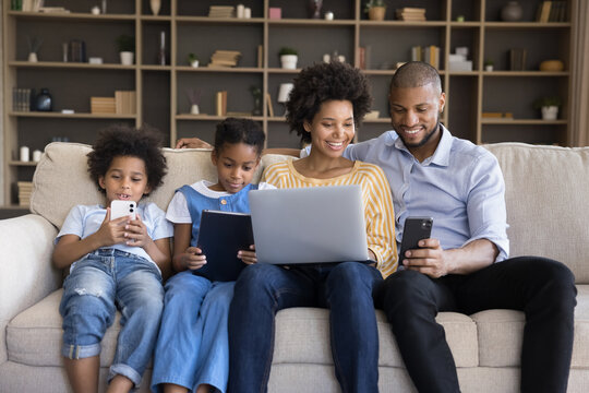 African spouses and children sit on sofa enjoy leisure use diverse electronic gadgets. Kids absorbed in videogame, instead live communication prefer online fun, bad life habit, devices overuse concept - Powered by Adobe