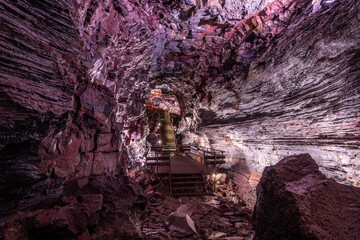 Ein Weg und eine Treppe durch lila beleuchtete Felsstrukturen im Lavatunnel Raufarholshellir auf...