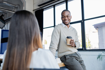Fototapeta premium smiling african american businessman holding paper cup and talking with blurred colleague.