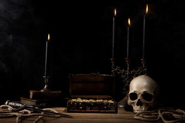 Old wooden treasure chest full of gold coins on the wooden table. Skull, dagger, candles,books and rope.