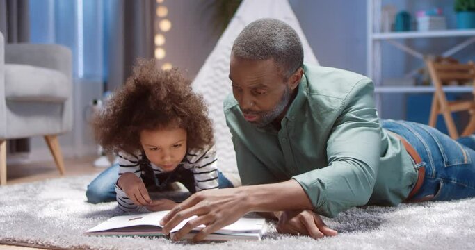 Close up of happy African American grandfather lying on floor with his little granddaughter and reading her children's book, looking at beautiful illustrations.