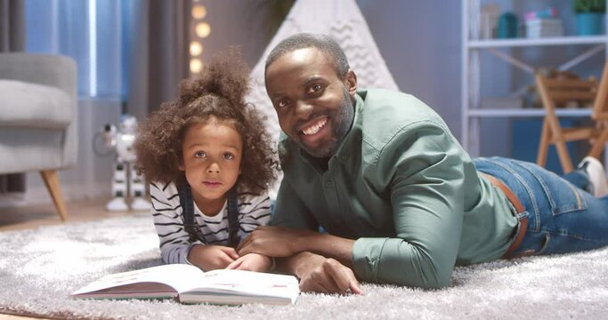 Portrait of happy african american grandfather and little girl with curly hair lying on floor with children's book and smiling at camera.