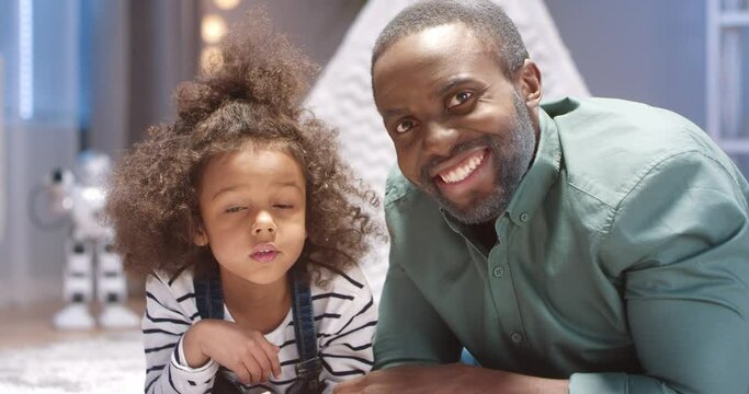 Close up portrait of happy african american grandfather and granddaughter with curly hair lying on floor with children's book and smiling at camera.