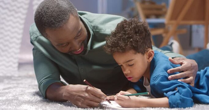 Close up of happy African American grandfather lying on floor with his little grandson and drawing picture together creating interesting story.