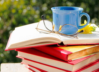 Knowledge and pleasure. Stack of books, a cup of coffee, glasses and flower on table outdoors in sunny day