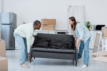 happy interracial couple carrying sofa in living room.