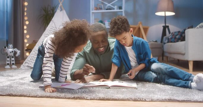 Cute African American brother and sister sitting on the floor at home and looking at new paper book while Grandpa tells stories.