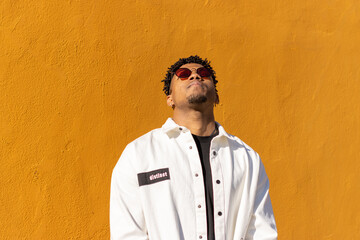 portrait of black latin man wearing sunglasses with closed eyes looking upwards on yellow background