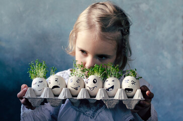 Little girl holding box with easter eggs with faces with fresh cress. Creative fun for child with...