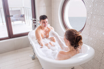 Young millennial woman taking a bath, having fun, making a white foam mustache on a mans face. A couple in love is having a fun weekend morning. The lovers kiss. Happy Newlyweds Honeymoon at the sea