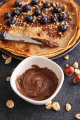 Bowl with tasty chocolate paste, nuts and crepes on black background