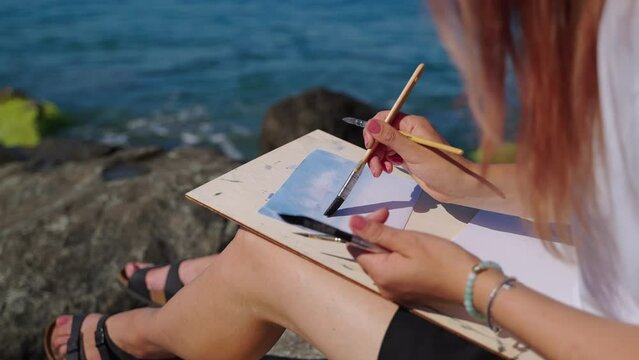 female student of school of art is painting marine landscape sitting on coast