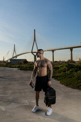 Chico joven musculado y tatuado posando sin camiseta y mochila delante de un puente