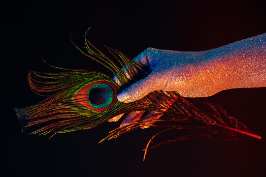 Peacock feather in female hand covered with holographic shining glitter under neon colored light. Body art, glamorous conceptual picture