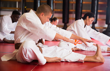 Caucasian man and group of people in kimono and belts doing stretch exercises on floor during karate training.. © JackF