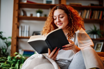 A beautiful ginger girl sitting in the living room wrapped in a blanket and reading an interesting book in the morning.