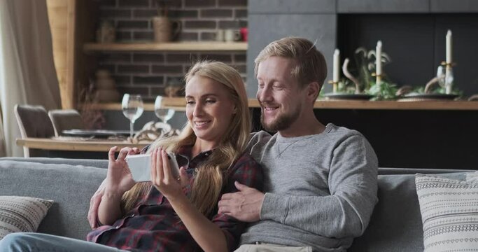 Happy blonde female and male enjoy weekend recreational home activity watching entertainment video smartphone. Funny casual couple choosing real estate for buying use helpful mobile phone application