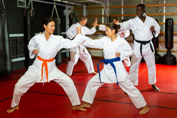 Fototapeta premium Man and women in kimono sparring together in gym during group karate training. African-american man trainer standing nearby and observing..