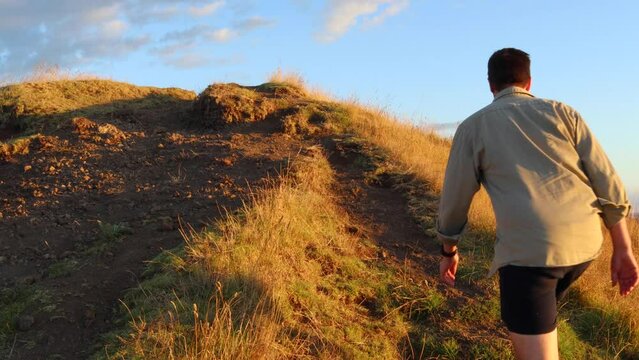 Young caucasian man in 30's walking up a dirt track on a grassy hill in the late afternoon