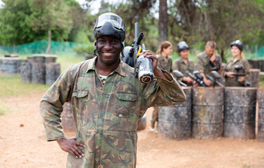 Fototapeta premium Smiling afro american man paintball player in camouflage posing with gun outdoors