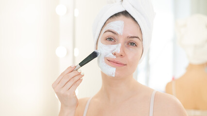 Beautiful caucasian woman with a towel on her hair applies a clay face mask. Taking care of beauty at home