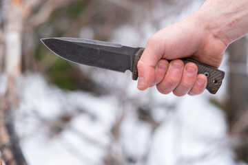 A close-up of a survival knife in a man's hand outdoors in a forest.