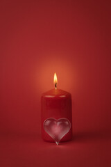 A small glass heart and a burning red candle on a red background.
