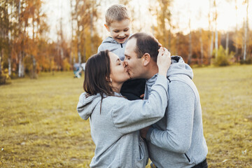 Fototapeta premium Mom and dad holding their son on their shoulders while they kiss and the child happily watches, spending happy time in the park. Portrait of parents kissing