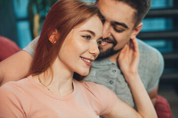 Close-up view of a Caucasian couple sitting on the couch on the weekend, freckled woman with red hair touching sensually her boyfriend's face.