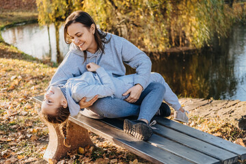 Mom sitting on the park bench holding her little girl lying on her arms laughing together. Family care. Happy family leisure. Smiling happy child.