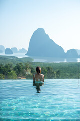 Happy woman in white swimsuit swimming in luxury pool hotel against Phang Nga bay background,...