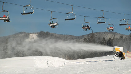 Snow cannon is working at ski resort the backdrop of snow-covered spruce forest, pine trees, ski slope on sunny winter morning. Chairlift moves, skiers sit on it. People ride on snowy mountainside