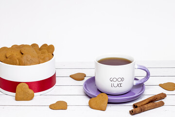 Ceramic cup with the inscription Good luck, heart-shaped cookies and cinnamon sticks on a light wooden background. Festive serving for a romantic breakfast.