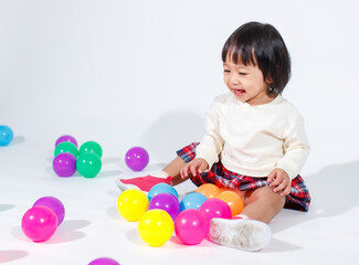 Studio shot of little cute short black hair Asian baby girl daughter model in casual plaid skirt sitting on floor smiling laughing playing with colorful round balls toy alone on white background