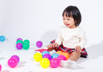 Fototapeta na wymiar Studio shot of little cute short black hair Asian baby girl daughter model in casual plaid skirt sitting on floor smiling laughing playing with colorful round balls toy alone on white background