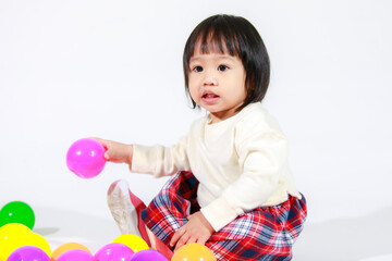 Studio shot of little cute short black hair Asian baby girl daughter model in casual plaid skirt sitting on floor smiling laughing playing with colorful round balls toy alone on white background