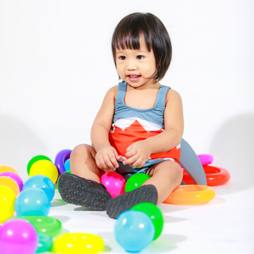 Studio shot of little beautiful kindergarten Asian baby girl daughter model in cute gray shark costume swimsuit outfit sitting on floor with colorful plastic balls and rings toy on white background