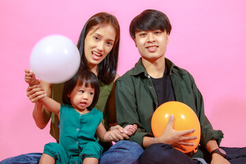 Fototapeta na wymiar Portrait closeup studio shot of young happy Asian family father mother and little cute baby girl daughter sitting on floor together smiling look at camera holding colorful balloons on pink background