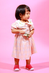 Portrait studio shot little cute Asian kindergarten preschooler girl daughter model in casual long dress and shoes standing look at camera holding hundred dollars banknote in hands on pink background