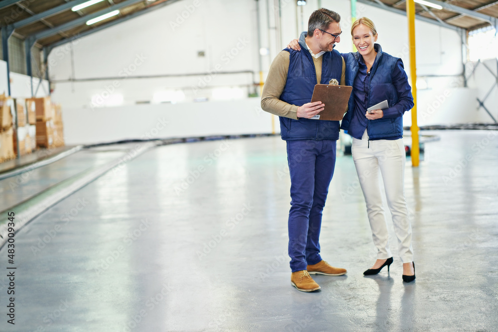 Poster comparing their inventory lists. shot of two coworkers talking while standing in a large warehouse.