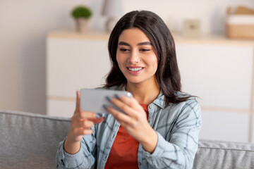 Lovely young Arab woman sitting on couch with smartphone, watching movie, working or studying from home