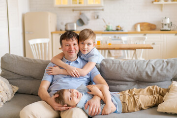 Happy caucasian father and little sons are having fun together, laughing dad hugs, tickling cute baby, sitting together on sofa at home in kitchen, enjoying the weekend, having fun together close-up